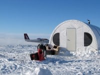 A small white shelter in the snow with a plane in the background