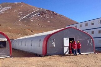 Four people in red jackets stand outside a long, gray and red tent in a mountainous area