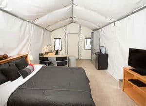 An interior view of a supervisor dormitory using a 12' wide x 20' long GB Series™ fabric building from Alaska Structures showcases ample space for a double bed, TV stand, small kitchenette, and a large workstation, ensuring a comfortable and functional living area.