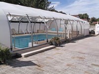 A swimming pool is covered by a fabric tent, with chairs and plants around it on a sunny day