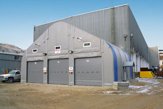 A large gray fabric tent with three roll-up doors next to a bigger industrial building