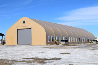 Large modular building with a roll-up door and multiple windows, set on a snowy ground under a clear blue sky