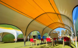 A sunny outdoor classroom setup with colorful tents, red chairs, and wooden desks
