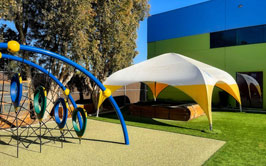 A colorful playground with a blue climbing structure and a white and yellow fabric tent on a grassy area