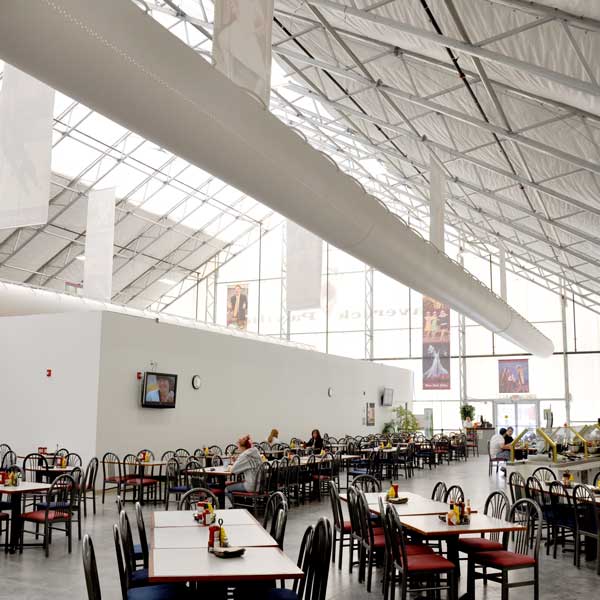 A spacious cafeteria inside a large, industrial-style fabric building with lots of tables and chairs under a high, bright ceiling