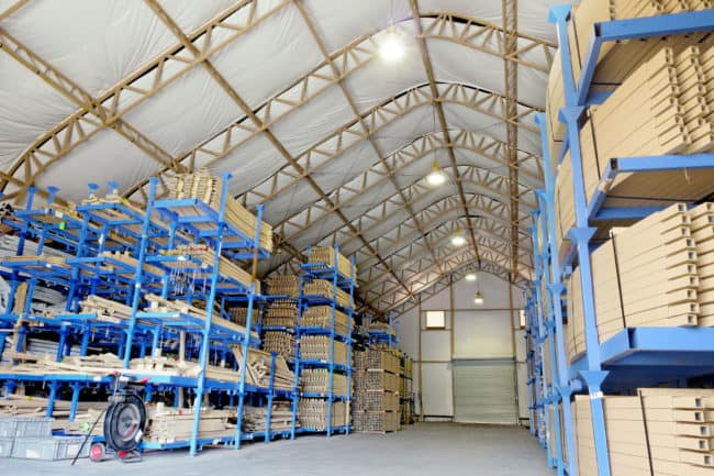 alaska-structures-industrial-warehouse-and-storage-building-1 A large, insulated DAGB Series™ fabric building from Alaska Structures® is anchored to a concrete slab and used to store powder-coated components for single-arch building solutions, featuring lighting, electrical systems, and large metal roll-up doors for easy access.