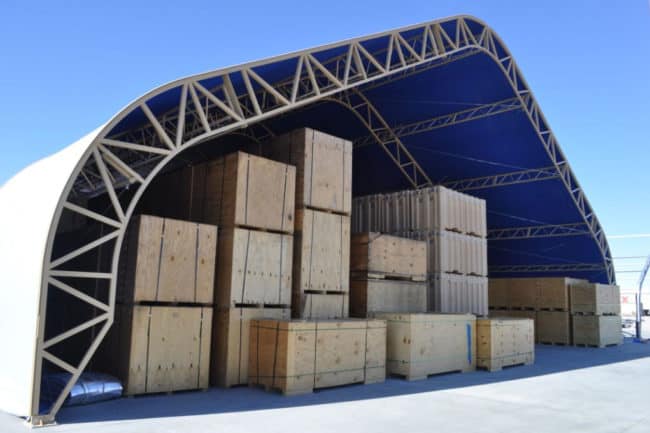 alaska-structures-industrial-warehouse-and-storage-building-9 A large open-ended warehouse, featuring a tan powder-coated frame DAGB Series™ fabric building from Alaska Structures®, is used to store wood and reusable shipping containers, providing protection from the elements.