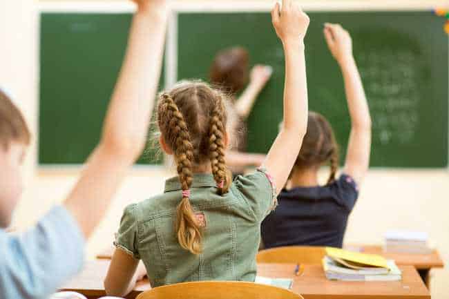 Girl with braids sitting at a desk raising her hand at the teacher writing on a black board with her back to the students.