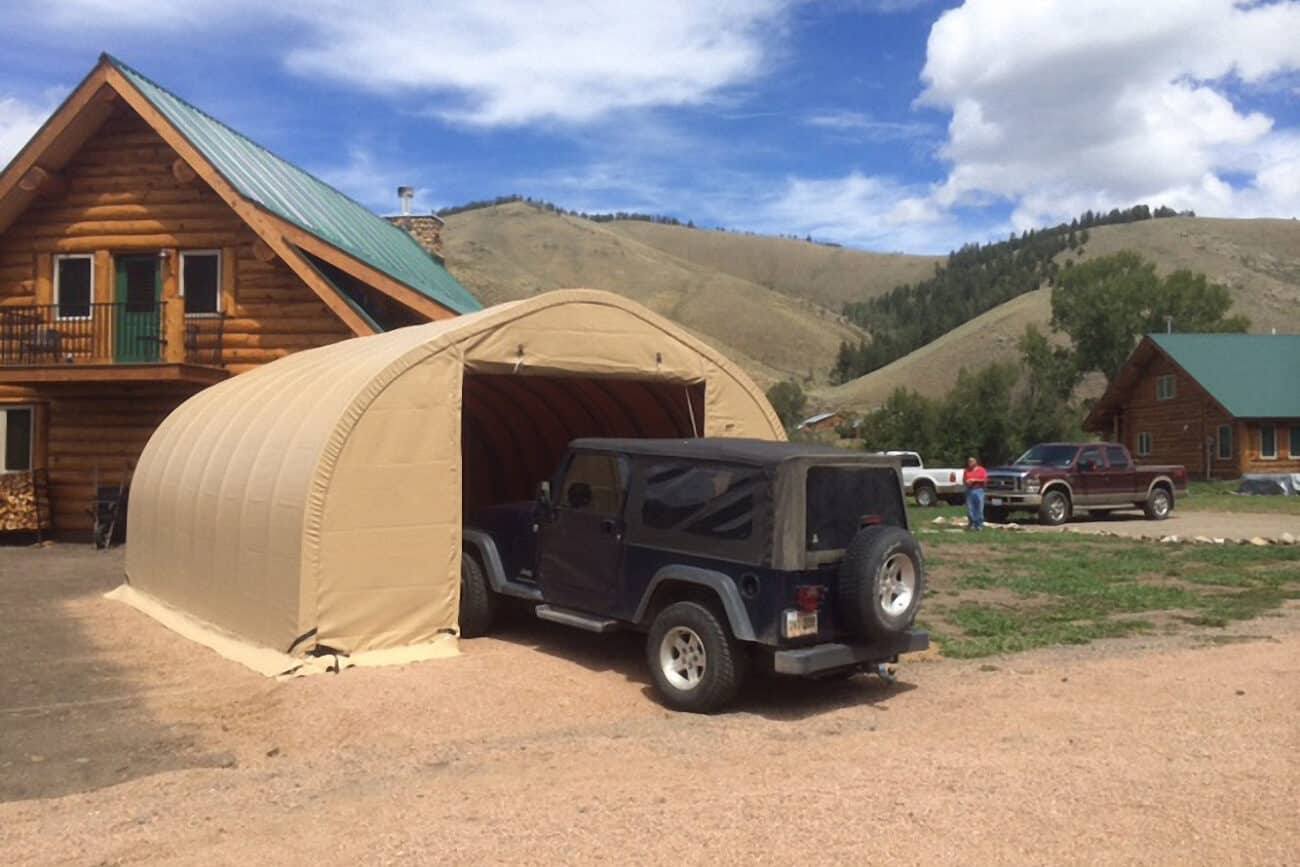 weatherport-portable-garage-hgb-series-6 A Jeep pulls into a tan WeatherPort® Portable Garage with a roll - up zipper door from Alaska Structures®, offering convenient vehicle storage at a log cabin resort.