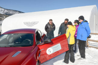A group of people checking out a red car by a large white fabric structure on a snowy day.
