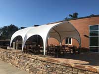A patio with tables and chairs is shaded by a white fabric canopy tent next to a building