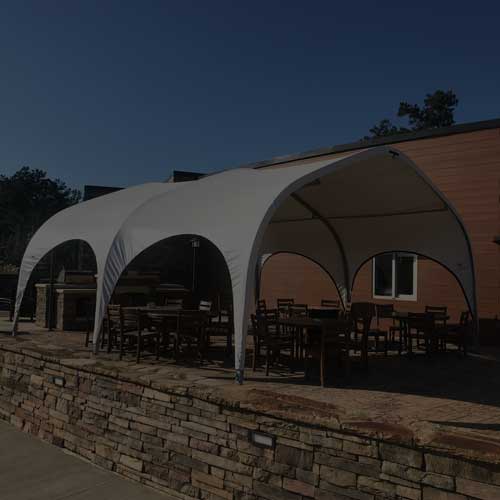 A patio with tables and chairs is shaded by a white fabric canopy tent next to a building